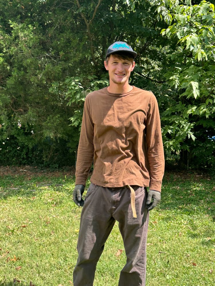 Amos Bear, owner of Mr Amos Landscaping, smiling in front of a mowed lawn and trimmed trees.